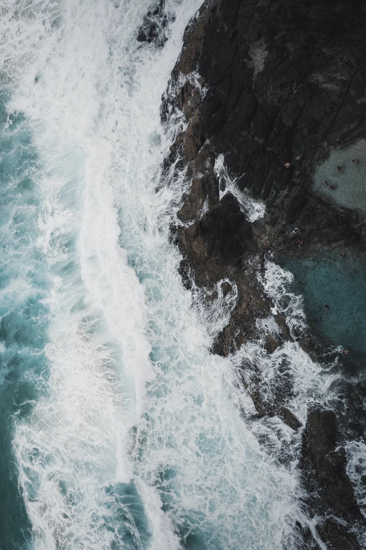 Surf meeting a dark rock shoreline, photographed from above.